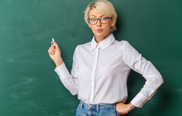 Strenge junge blonde Lehrerin mit Brille im Klassenzimmer, die vor der Tafel steht und auf die Tafel zeigt, wobei Kreide die Hand auf der Taille hält und nach vorne schaut