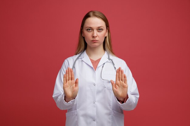 Strenge junge blonde Ärztin, die medizinische Robe und Stethoskop um den Hals trägt und Stoppgeste tut