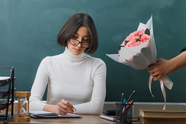 Strenge junge Lehrerin, die mit Schulwerkzeugen am Schreibtisch saß, erhielt im Klassenzimmer einen Blumenstrauß