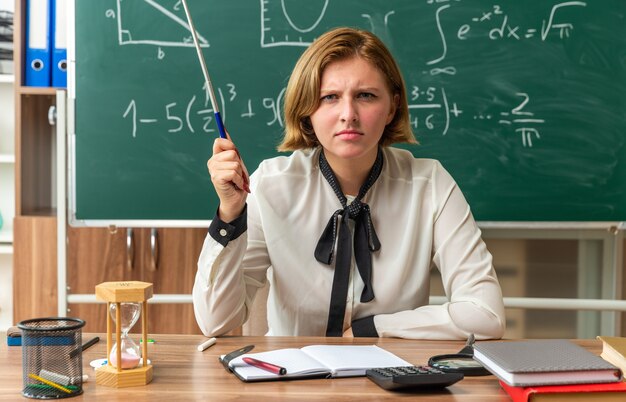 Strenge junge Lehrerin sitzt am Tisch mit Schulmaterial, das einen Zeigerstock im Klassenzimmer hält