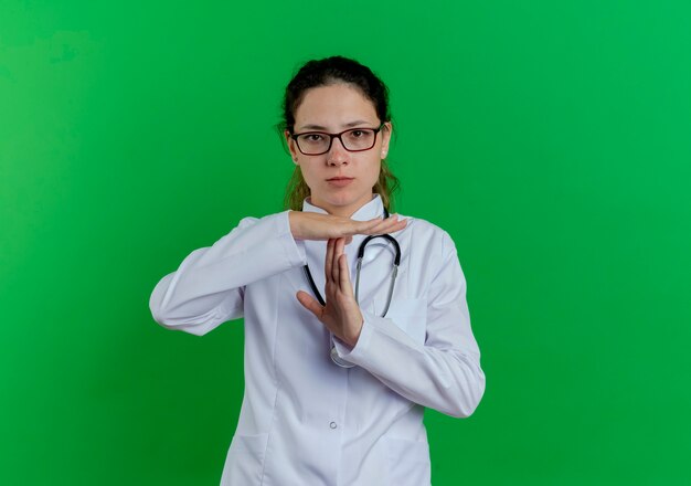 Strenge junge Ärztin, die medizinische Robe und Stethoskop und Brille trägt, lokalisiert, die Zeitüberschreitungsgeste tut, lokalisiert auf grüner Wand mit Kopienraum