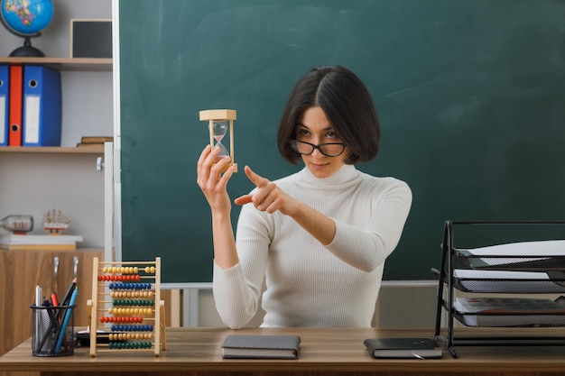 Strenge Punkte an der Seite Junge Lehrerin mit Brille, die Sanduhr hält, sitzt am Schreibtisch mit eingeschalteten Schulwerkzeugen im Klassenzimmer