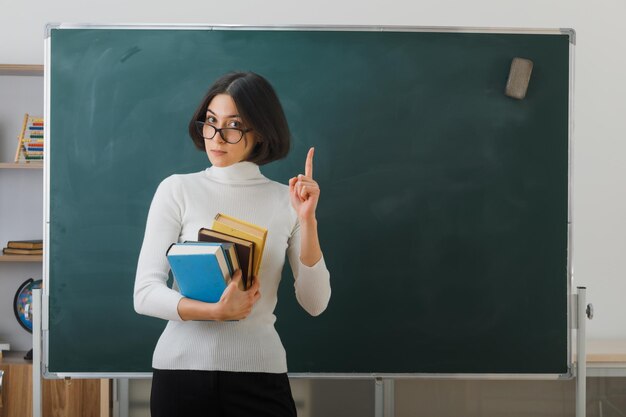 Strenge Punkte auf junge Lehrerin mit Brille, die Bücher hält, die vor der Tafel im Klassenzimmer stehen