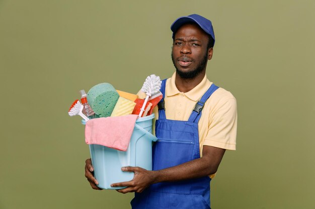 Strenger Halteeimer mit Reinigungswerkzeugen junger afroamerikanischer Reinigungsmann in Uniform mit Handschuhen isoliert auf grünem Hintergrund