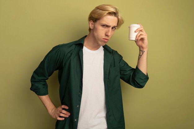 Strenger junger blonder Kerl, der grünes T-Shirt trägt, das Tasse Kaffee hält und Hand auf Hüfte legt