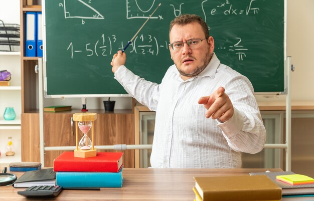 Strenger junger Lehrer mit Brille, der am Schreibtisch mit Schulmaterial im Klassenzimmer sitzt und auf die Vorderseite zeigt, die auf die Tafel zeigt, mit dem Zeigerstock, der nach vorne schaut und zeigt