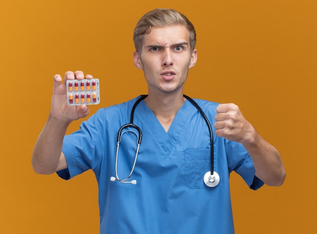 Strenger junger männlicher Arzt, der Arztuniform mit Stethoskop hält, das Pillen hält, die auf orange Wand lokalisiert werden