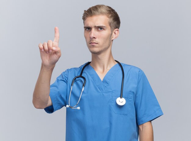 Strenger junger männlicher Arzt, der Arztuniform mit Stethoskopspitzen oben auf weißer Wand trägt