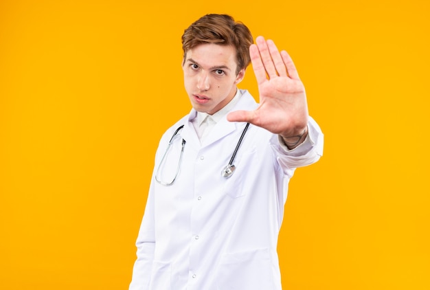Strenger junger männlicher Arzt, der ein medizinisches Gewand mit Stethoskop trägt, das die Stoppgeste isoliert auf der orangefarbenen Wand zeigt