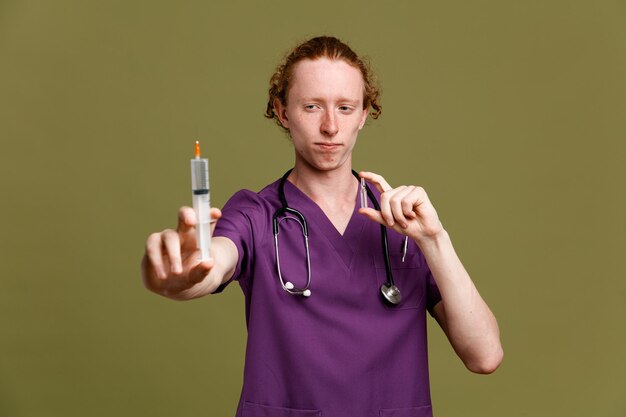 Strenger junger männlicher Arzt in Uniform mit Stethoskop mit Spritze mit Ampulle isoliert auf grünem Hintergrund