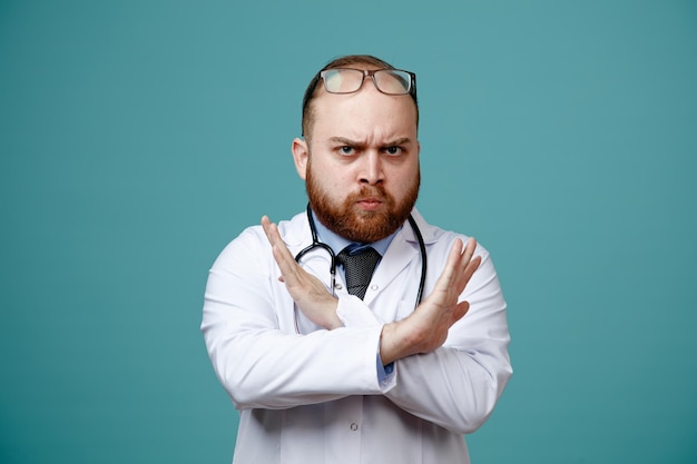 Strenger junger männlicher Arzt mit Arztkittel und Stethoskop um den Hals und Brille auf dem Kopf, der in die Kamera blickt und keine Geste isoliert auf blauem Hintergrund zeigt