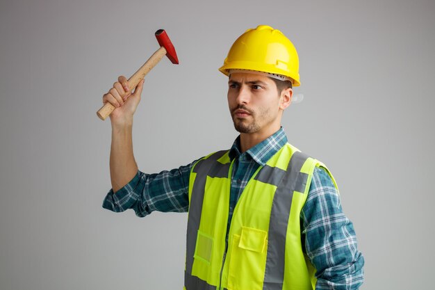 Strenger junger männlicher Ingenieur mit Schutzhelm und Uniform, der in der Profilansicht steht und den seitlich anhebenden Hammer isoliert auf weißem Hintergrund betrachtet