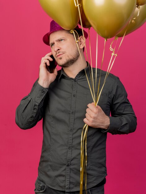 Strenger junger Party-Typ, der rosa Hut hält, der Luftballons hält und am Telefon isoliert auf rosa spricht