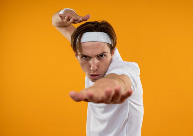 Strenger junger sportlicher Kerl, der Stirnband und Armband trägt, die Hand lokalisiert auf orange Wand halten
