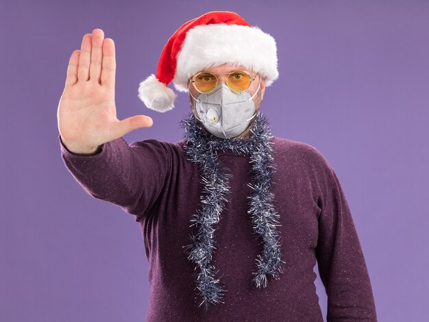 Strenger Mann mittleren Alters, der Weihnachtsmütze und Schutzmaske mit Lametta-Girlande um den Hals mit Brille trägt und Stoppgeste tut, die auf lila Wand isoliert wird
