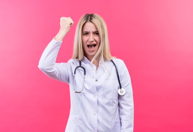 Strenges junges blondes Mädchen des Arztes, das Stethoskop im medizinischen Kleid hält, das Hand auf lokalisiertem rosa Hintergrund hält