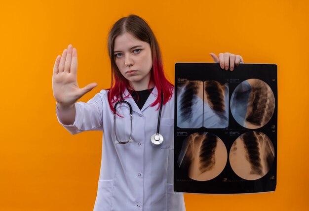 Strenges junges Doktormädchen, das medizinisches Gewand des Stethoskops trägt, das Röntgenbild hält, das Stoppgeste auf lokalisiertem orangefarbenem Hintergrund zeigt