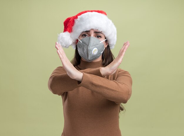 Strenges junges schönes Mädchen, das Weihnachtsmütze mit medizinischer Maske trägt, die Geste von nicht isoliert auf olivgrünem Hintergrund zeigt