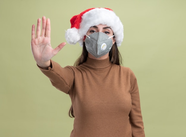 Strenges junges schönes Mädchen, das Weihnachtsmütze mit medizinischer Maske trägt, die Stoppgeste zeigt, die auf olivgrünem Hintergrund lokalisiert wird