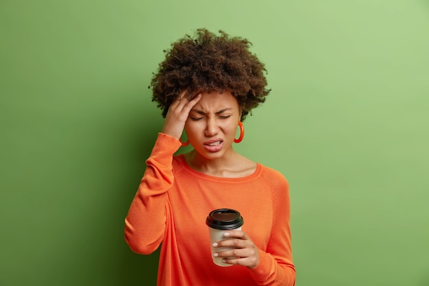 Stressvolle afroamerikanische Frau fühlt schreckliche Kopfschmerzen schmunzelt vor Schmerz schließt Augen berührt Schläfengetränke Kaffee trägt lässigen orangefarbenen Pullover, der über leuchtend grüner Wand isoliert ist