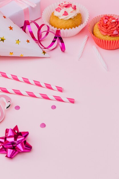 Strohhälme; Bandschleife; Partyhut; Luftschlangen; Geschenkbox; Cupcake und Kerzen auf rosa Hintergrund