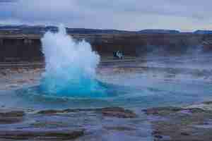 Kostenloses Foto strokkur geysir, umgeben von hügeln unter bewölktem himmel am abend in island