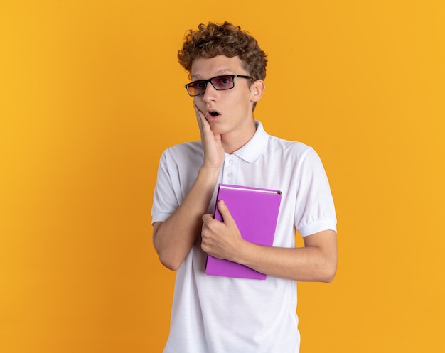 Student in Freizeitkleidung mit Brille, die ein Buch hält und die Kamera anschaut, erstaunt und überrascht, über orangefarbenem Hintergrund zu stehen