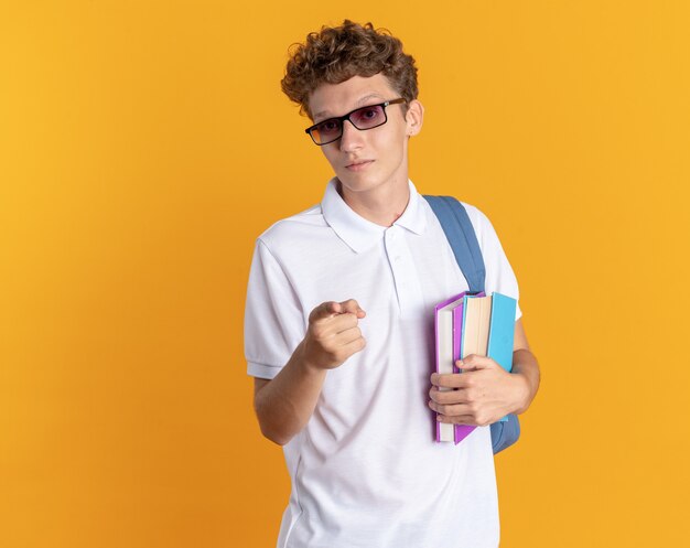 Student in Freizeitkleidung mit Brille mit Rucksack, der Bücher hält, die selbstbewusst mit dem Zeigefinger auf die Kamera zeigt, die über orangefarbenem Hintergrund steht
