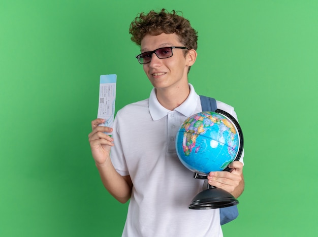 Student in Freizeitkleidung mit Brille mit Rucksack, der Globus und Flugticket hält und selbstbewusst in die Kamera schaut
