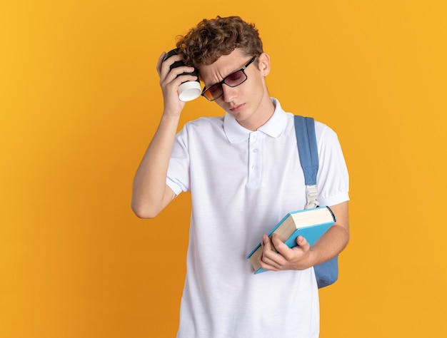 Student in Freizeitkleidung mit Brille mit Rucksack mit Buch und Pappbecher, der müde aussieht looking