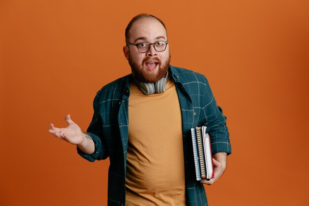 Student in Freizeitkleidung mit Brille und Kopfhörern, die Notizbücher in der Hand halten und verwirrt und überrascht den Arm über orangefarbenem Hintergrund heben