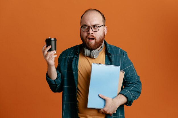 Student in Freizeitkleidung mit Brille und Kopfhörern mit Notizbüchern und Kaffeetasse, die verwirrt beiseite schaut und Zweifel hat, die über orangefarbenem Hintergrund stehen