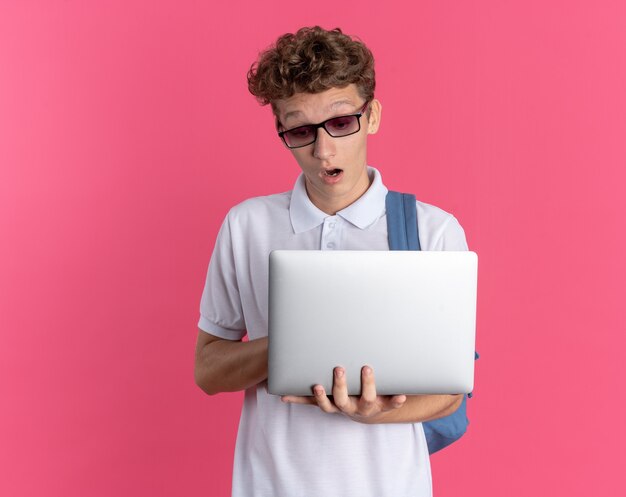 Student in Freizeitkleidung mit Brille und Rucksack mit Laptop, der überrascht und erstaunt auf rosafarbenem Hintergrund steht