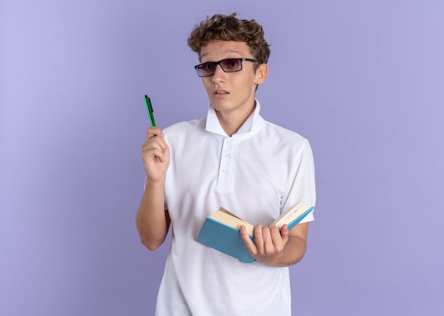 Student in weißem Poloshirt mit Brille mit Buch und Stift und Blick in die Kamera überrascht mit einer neuen Idee auf blauem Hintergrund