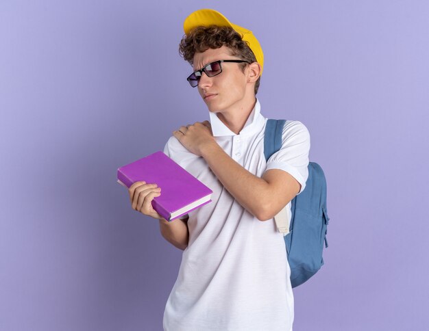 Student in weißem Poloshirt und gelber Mütze mit Brille mit Rucksack, der ein Notebook hält, das seine Schulter berührt und Schmerzen fühlt