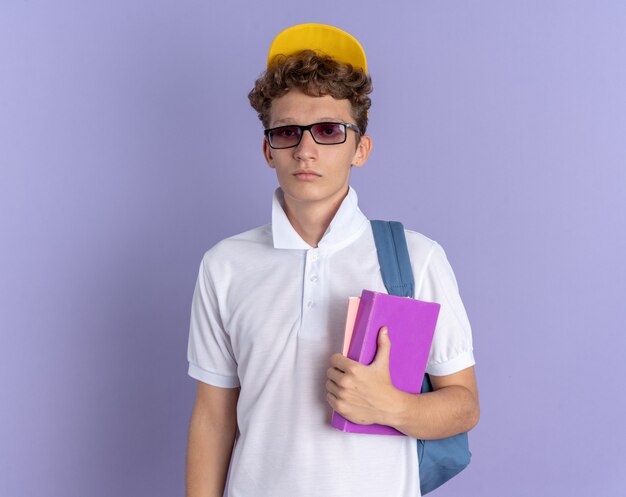 Student in weißem Poloshirt und gelber Mütze mit Brille mit Rucksack, der Notizbücher hält und mit ernstem Gesicht in die Kamera schaut