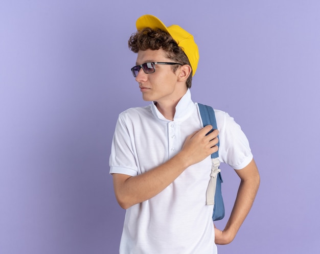 Student in weißem Poloshirt und gelber Mütze mit Brille und Rucksack, der mit ernstem Gesicht beiseite schaut