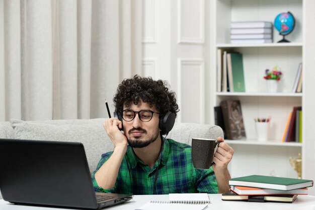 Student online süßer junger Kerl, der am Computer mit Brille im grünen Hemd studiert, müde denkend