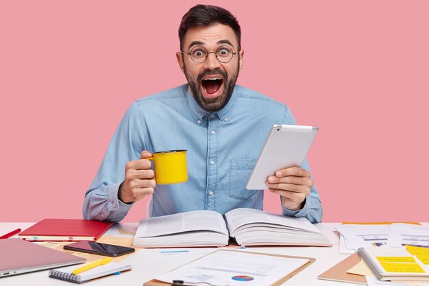 Student sitzt am Schreibtisch mit Dokumenten, die Tasse und Tablette halten