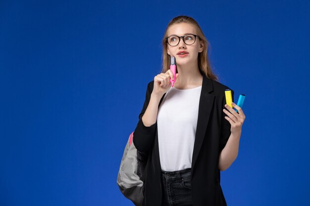 Studentin der Vorderansicht in der schwarzen Jacke, die den Rucksack hält, der Filzstifte hält, die auf blauen Wandstunden School College Universität denken