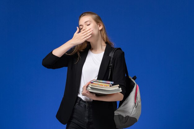 Studentin der Vorderansicht in der schwarzen Jacke, die Rucksack hält Bücher hält und auf der blauen Wandschule Universitätsuniversitätsstunde gähnt