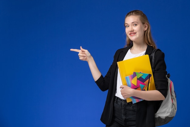Studentin der Vorderansicht in der schwarzen Jacke, die Rucksack hält Datei mit Heften auf blauen Wand College-Universitätsstunden trägt