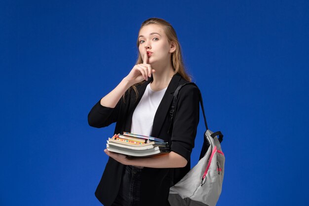 Studentin der Vorderansicht in der schwarzen Jacke, die Rucksack hält, der Bücher auf der Universitätsuniversitätsstunde der blauen Schreibtischschule hält