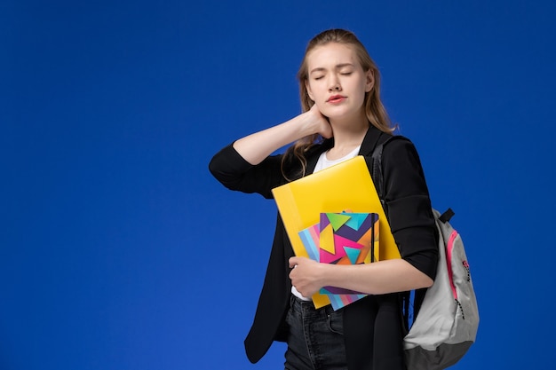 Studentin der Vorderansicht in der schwarzen Jacke, die Rucksack hält, der Dateien mit Heften hält, die Nackenschmerzen auf der Universitätslektion der blauen Wanduniversität haben