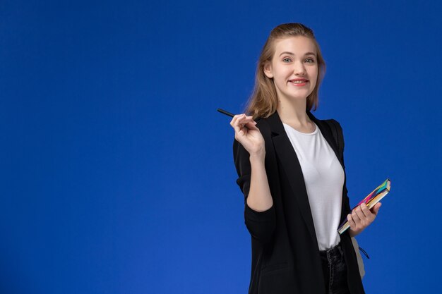 Studentin der Vorderansicht in der schwarzen Jacke, die Rucksack hält, der Stift und Heft hält, die auf Universitätslektion der blauen Wanduniversität lächeln