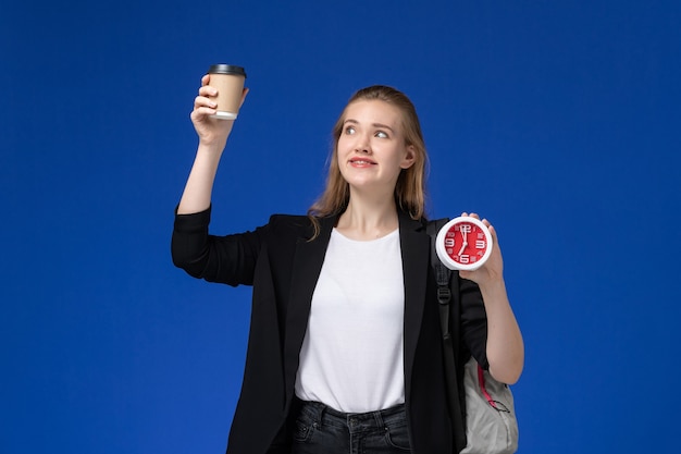 Studentin der Vorderansicht in der schwarzen Jacke, die Rucksack hält, der Uhren und Kaffee auf der blauen Wandschule College-Universitätsstunde hält