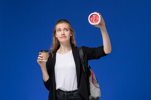 Studentin der Vorderansicht in der schwarzen Jacke, die Rucksack hält, der Uhren und Kaffee auf hellblauen Wandschuluniversitätsstunden hält