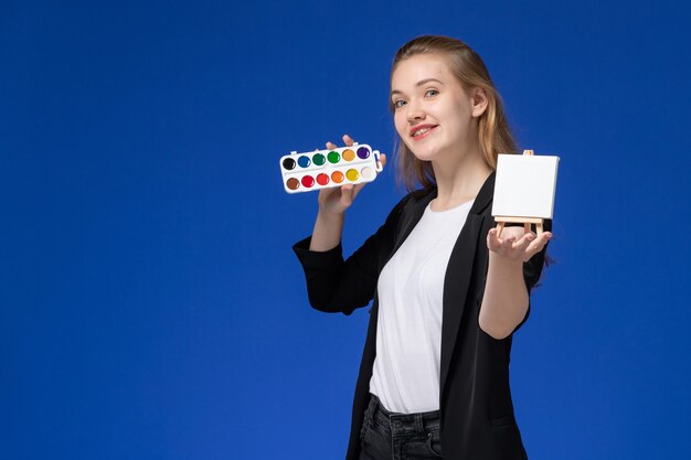 Studentin der Vorderansicht in der schwarzen Jacke, die Rucksack hält Farben für Zeichnung und Staffelei auf blauem Schreibtisch zeichnet Kunstschulhochschule