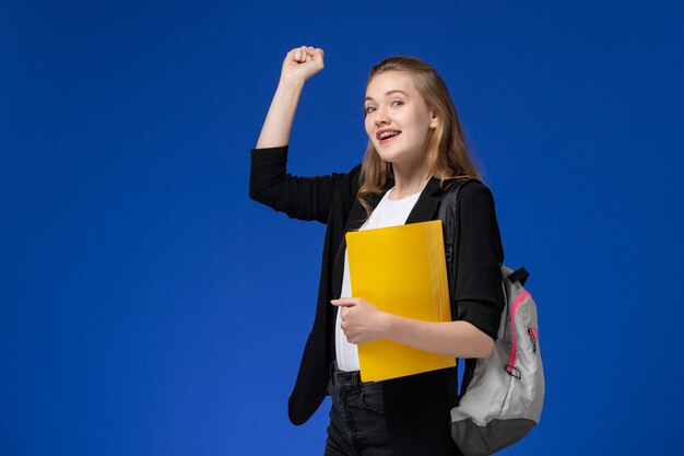 Studentin der Vorderansicht in der schwarzen Jacke, die Rucksack hält, gelbe Akten auf der blauen Schreibtischschule College-Universitätsstunde hält