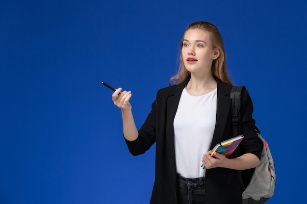 Studentin der Vorderansicht in der schwarzen Jacke, die Rucksack hält Stift und Heft auf hellblauer Wanduniversitätsstunde hält